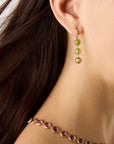 18K yellow gold drop earrings featuring bezel-set, rose-cut round peridot gemstones arranged in a continuous line with hook posts set with diamonds. Displayed on a model.