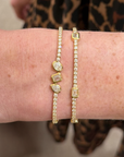 Two gold bracelets worn on a person's wrist, one with small emerald cut diamonds in a station style tennis bracelet. Along with a tennis bracelet featuring a pear shaped, emerald cut, and oval shaped diamond set in a bezel station on a tennis bracelet.