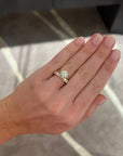 A close-up of a person's hand wearing a a round diamond solitaire engagement ring with a round diamond band ring, set against an indoor background with soft gray and beige tones