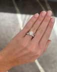 A close-up of a person's hand wearing a round solitaire diamond ring set in a white gold setting and paired with a medium white gold diamond wedding band, set with round diamonds. Set against a blurred indoor background.