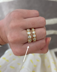 A close-up of a hand wearing two gold eternity rings set with clear round gemstones. A white arrow points to the rings, which are shown against a gray and white striped background with a white skirt featuring yellow dots.