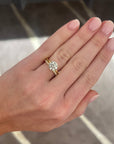 A close-up of a hand wearing a yellow gold ring with a large round-cut diamond cluster, set against a blurred gray patterned background.