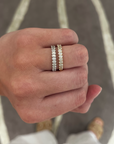 A close-up of a person's hand wearing two slim eternity rings. One ring is silver with clear stones, and the other is yellow gold with clear stones, set against a gray and white patterned rug.