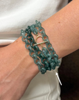 A close-up of a person's wrist wearing a multi-strand teal chain bracelet with a gold toggle clasp, against a white shirt background.