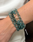 A close-up of a person's wrist wearing a multi-strand teal-green chain bracelet with a gold toggle clasp, against a white shirt background.