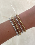 Four beaded stretch bracelets in silver and gold tones stacked on a person's wrist, with a white arrow pointing to the stack. The background is a soft beige marble-like surface.