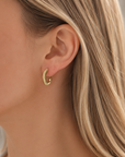 A close-up of a woman's ear wearing a small textured gold huggie hoop earring, with blonde hair visible against a soft neutral background.