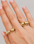 A close-up of a hand wearing two gold rings with a textured, ribbed beaded design, set against a plain light gray background.