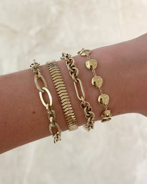 A close-up of a person's wrist wearing four gold-toned bracelets, including a snake chain, a link chain, a disc charm bracelet, and a textured chain.