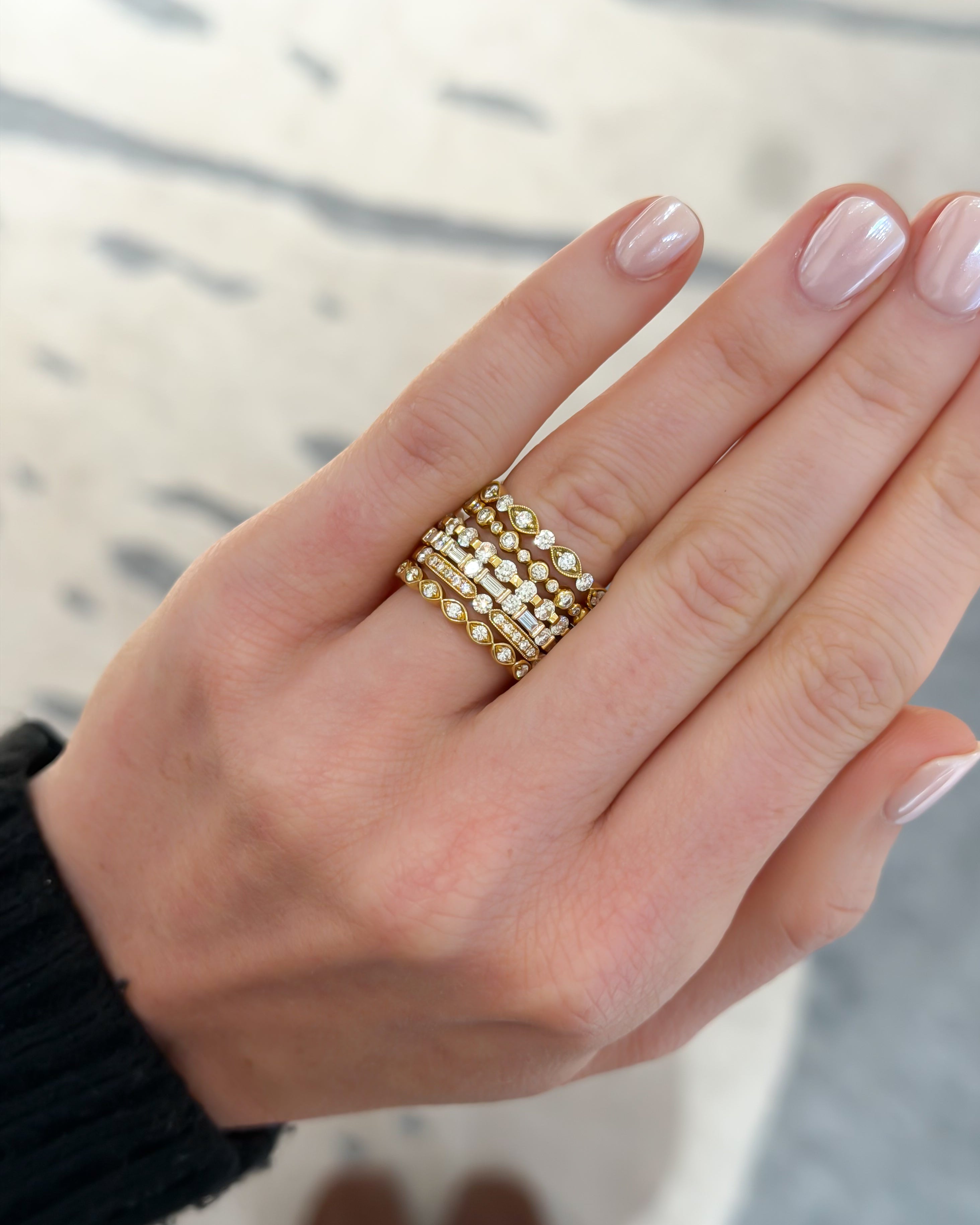 A close-up of a hand wearing a stack of three gold rings, each set with small clear stones, against a softly blurred light background.