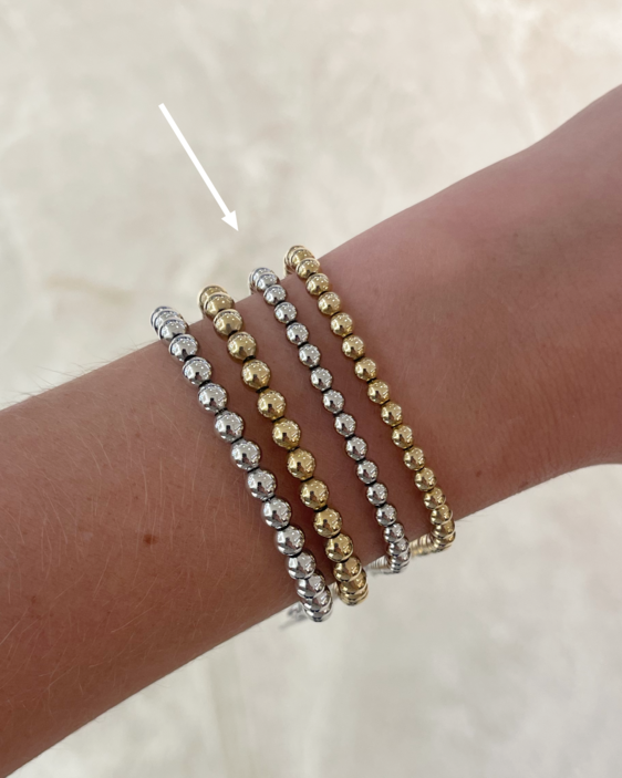 Photo of four beaded bracelets on a model's arm with an arrow pointing to a 14k white gold bracelet made of small beads that are strung together to a 7.5 inch bracelet with adjustable chain clasp.