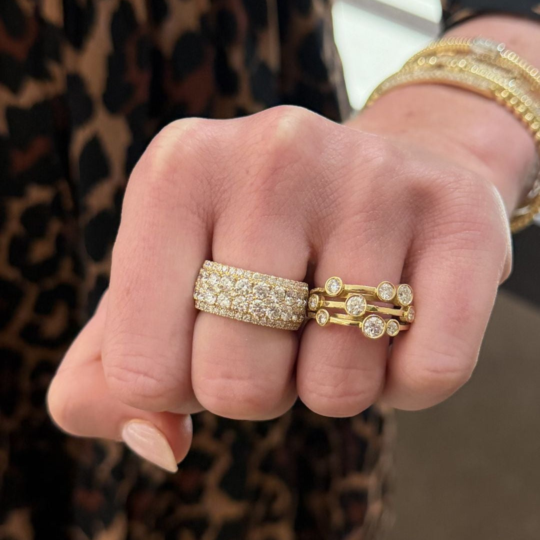 A close-up of a person's hand wearing two gold rings with pavé stones. One 3 row bezel set diamond band scattered round diamonds. 