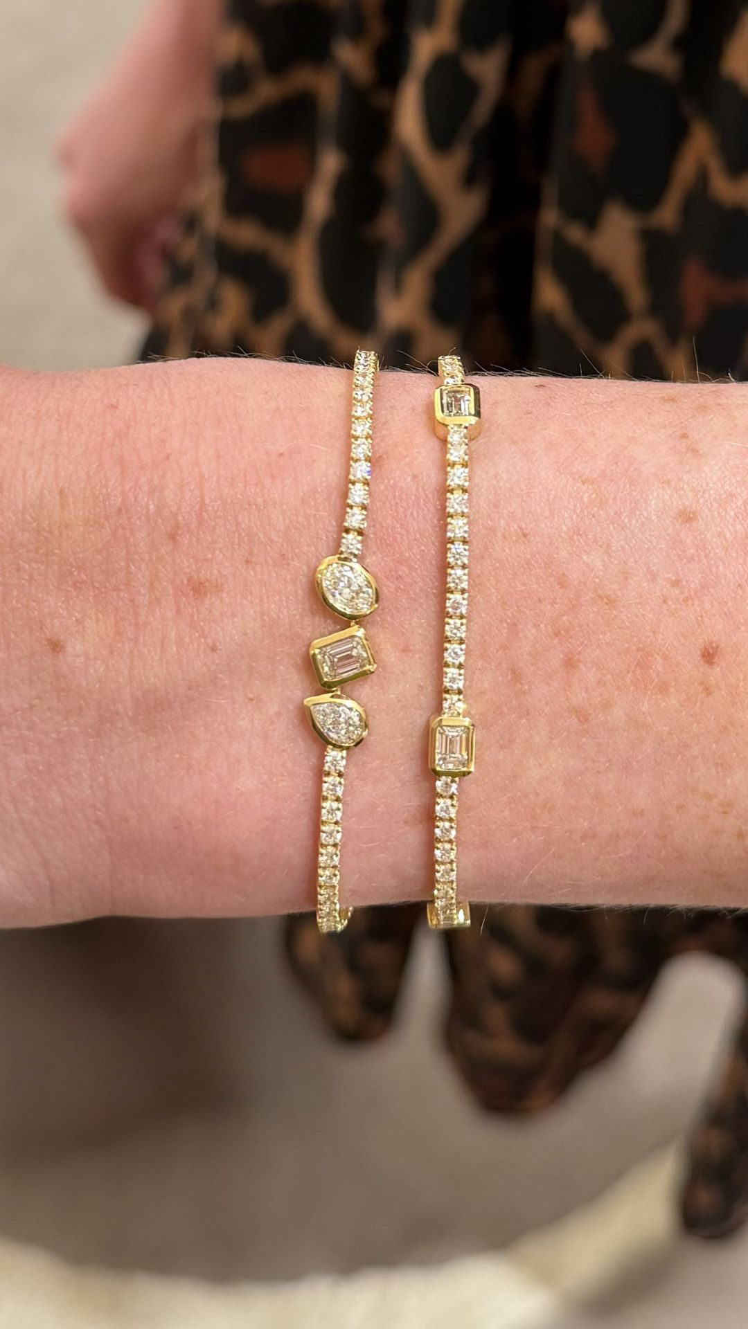 Two gold bracelets worn on a person's wrist, one with small emerald cut diamonds in a station style tennis bracelet. Along with a tennis bracelet featuring a pear shaped, emerald cut, and oval shaped diamond set in a bezel station on a tennis bracelet. 