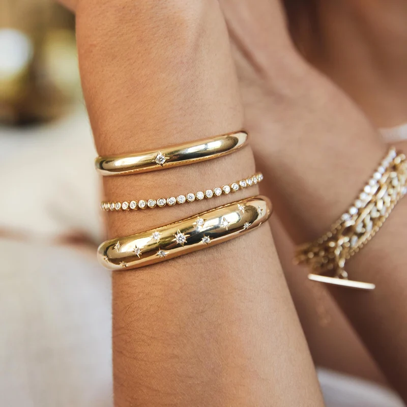 14k yellow gold cuff bracelet from Zoë Chicco features a single star-set diamond that totals 0.10ctw and at the center of the slim cuff. Shown and styled on a model's wrist stack.