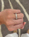 A close-up of a hand wearing a silver band ring with a textured, segmented design, set against a background of a gray patterned floor and white clothing.
