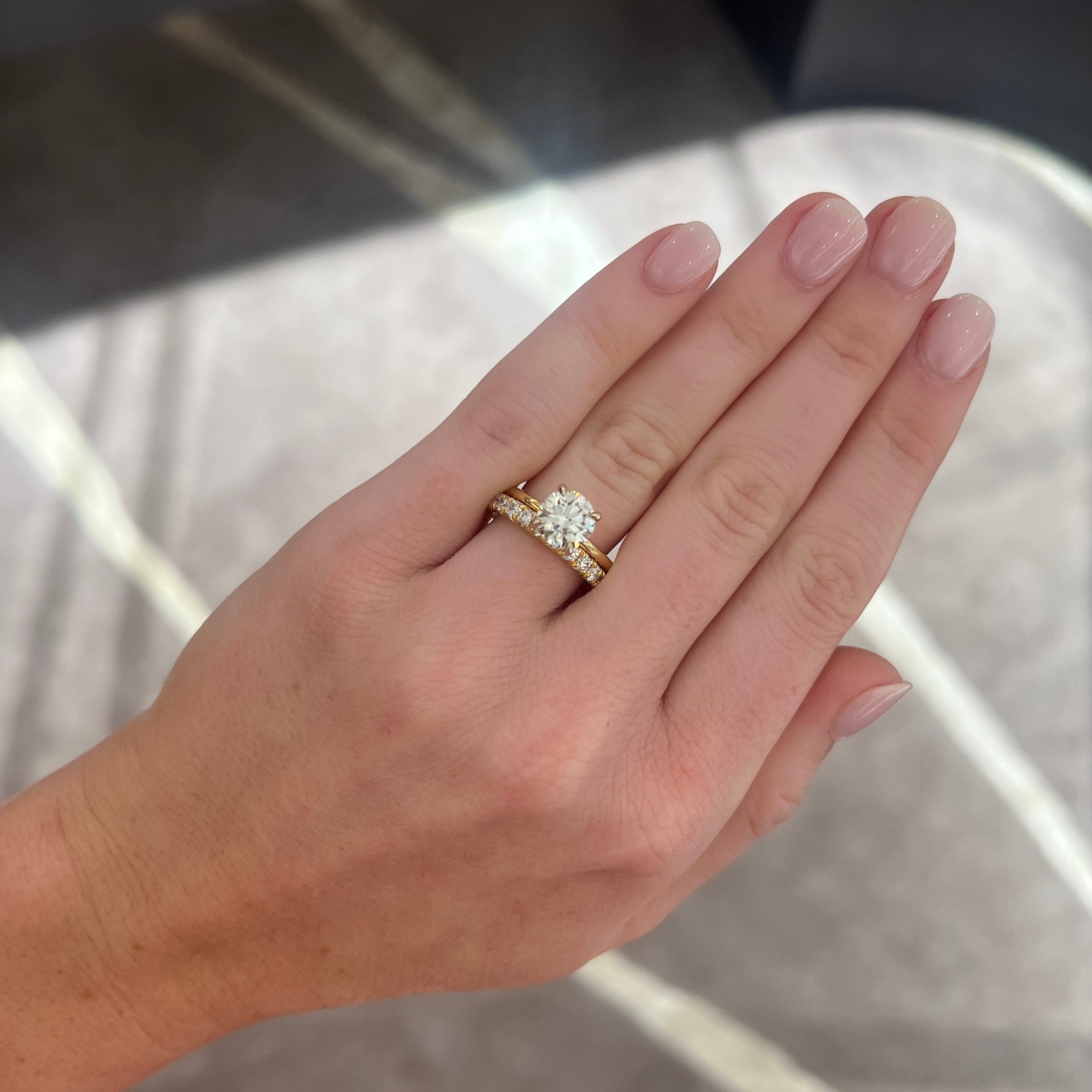 A close-up of a hand wearing a yellow gold ring with a round-cut diamond solitaire, paired with a yellow gold diamond band, set against a gray patterned background.