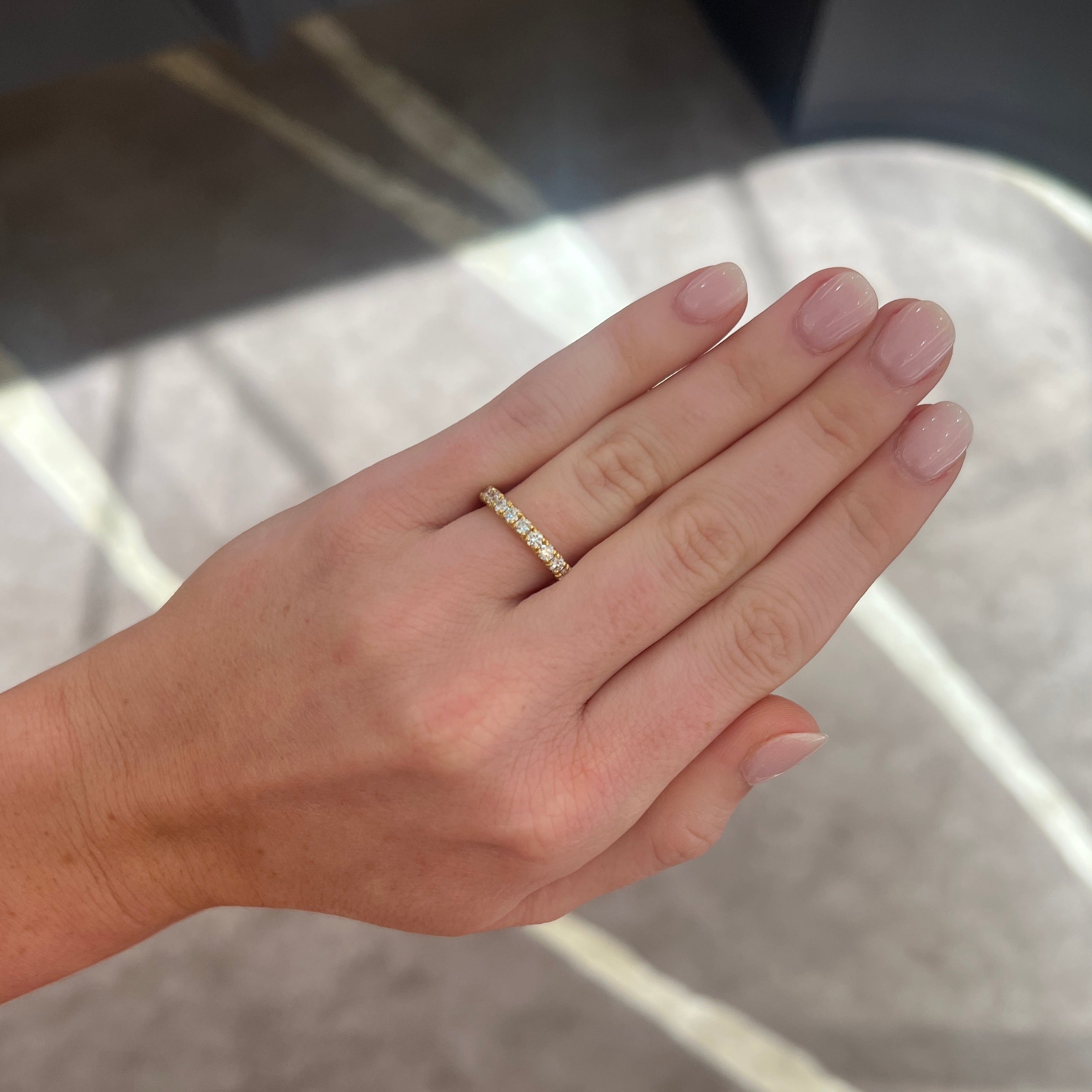 A close-up of a person's hand wearing a delicate gold band ring with round diamonds, set against a gray patterned background.