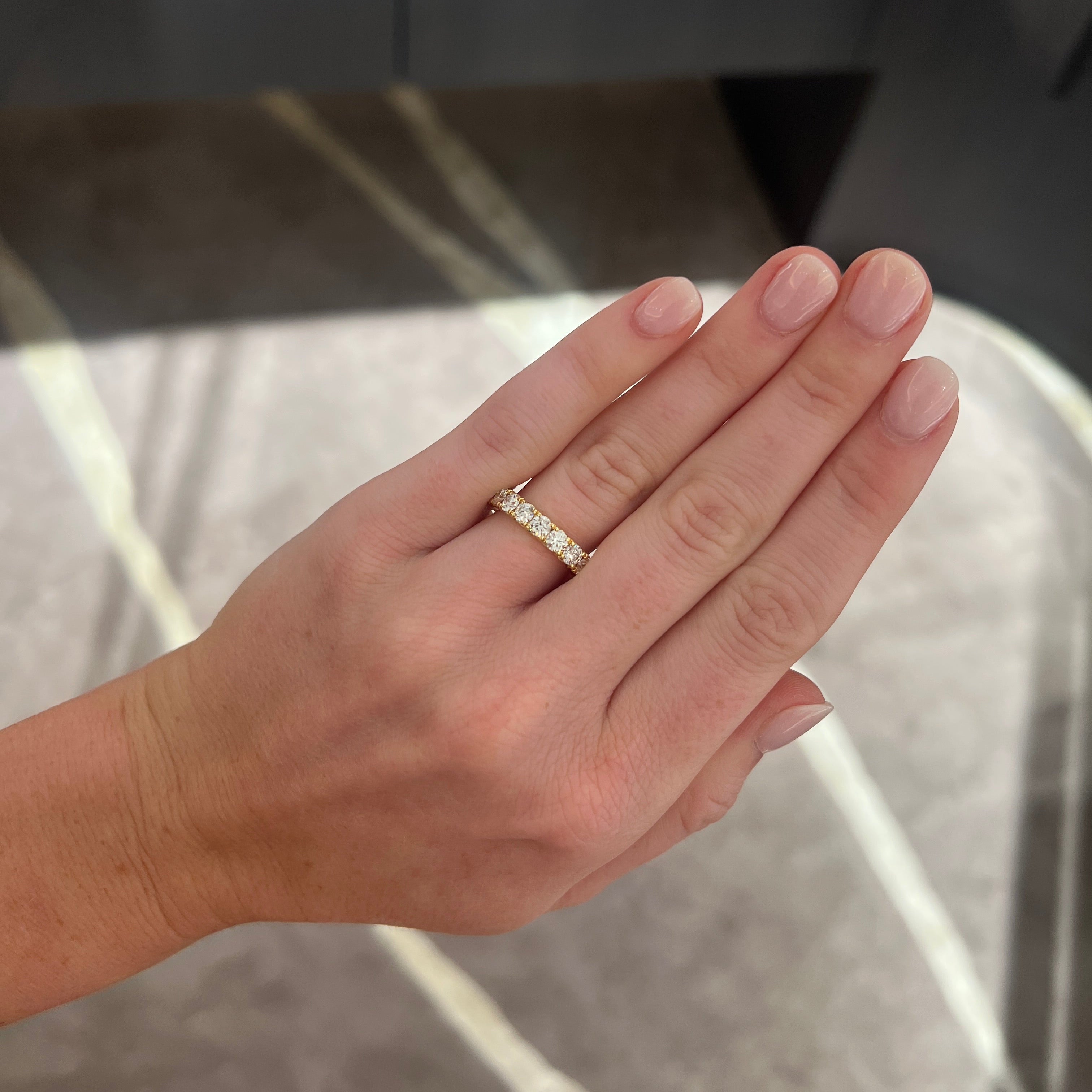 A close-up of a person's hand wearing a large round diamond band ring, set against an indoor background with soft gray and beige tones