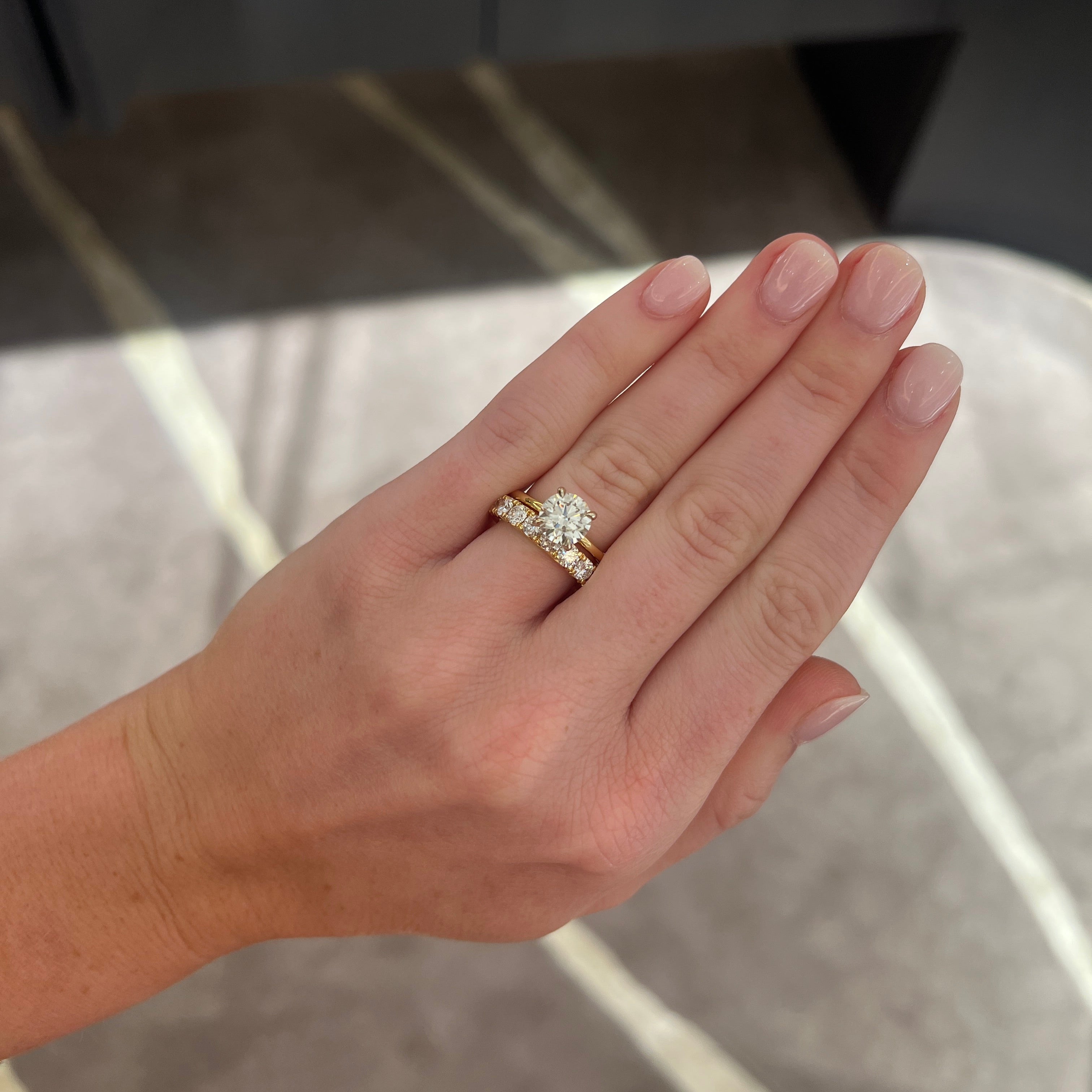 A close-up of a person's hand wearing a a round diamond solitaire engagement ring with a round diamond band ring, set against an indoor background with soft gray and beige tones
