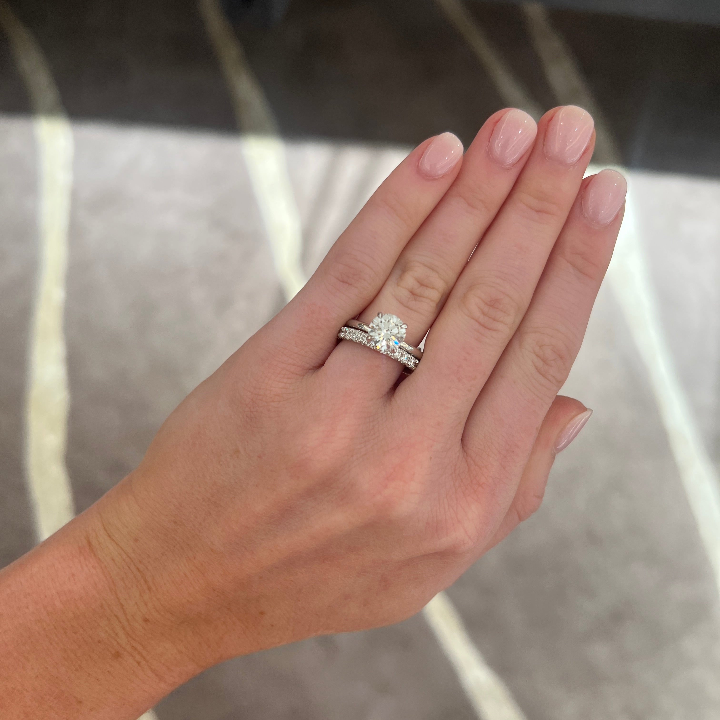 A close-up of a person's hand wearing a round solitaire diamond ring set in a white gold setting and paired with a medium white gold diamond wedding band, set with round diamonds. Set against a blurred indoor background.