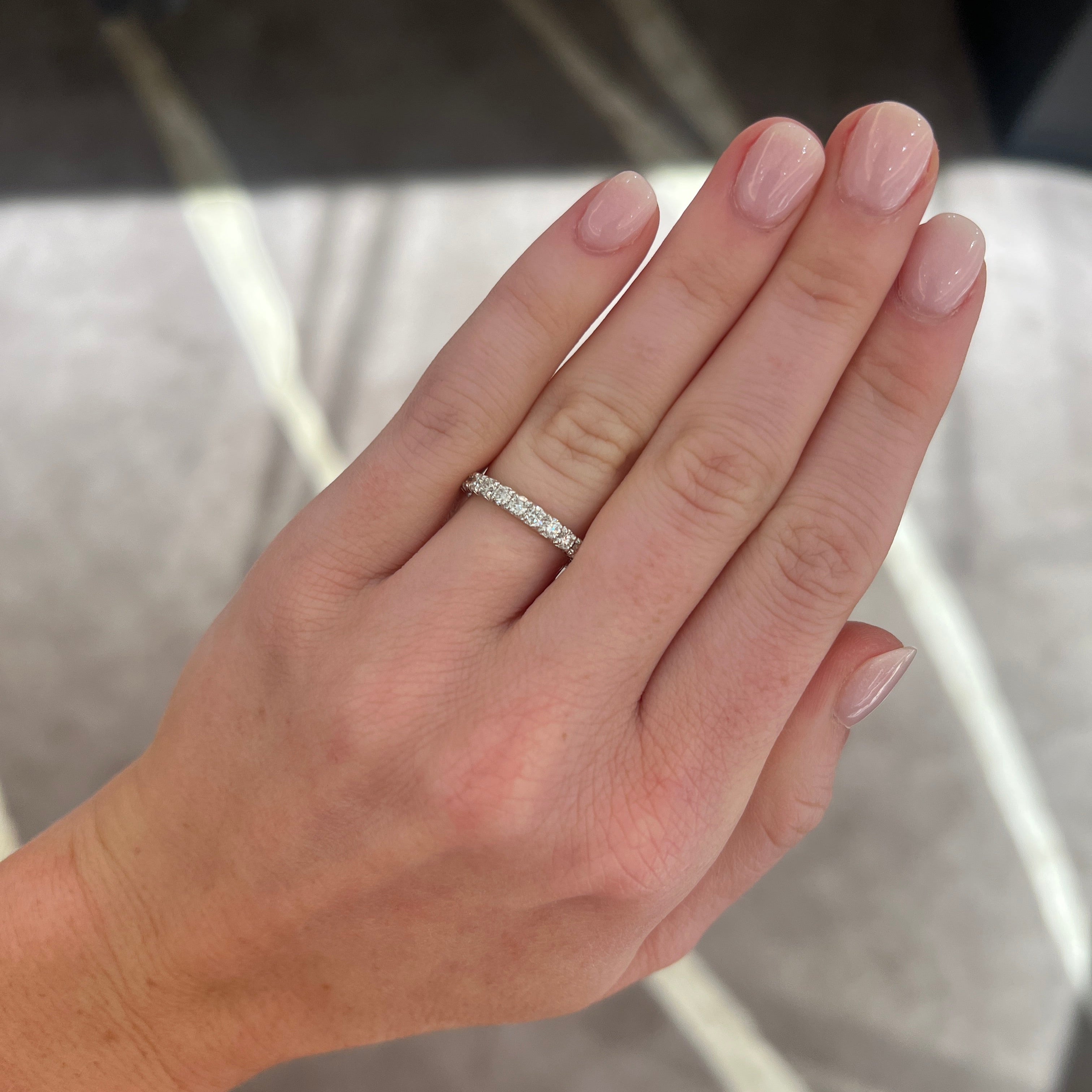 A close-up of a person's hand wearing a medium white gold diamond wedding band, set with round diamonds, set against a blurred indoor background.