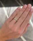 A close-up of a person's hand wearing a medium white gold diamond wedding band, set with round diamonds, set against a blurred indoor background.