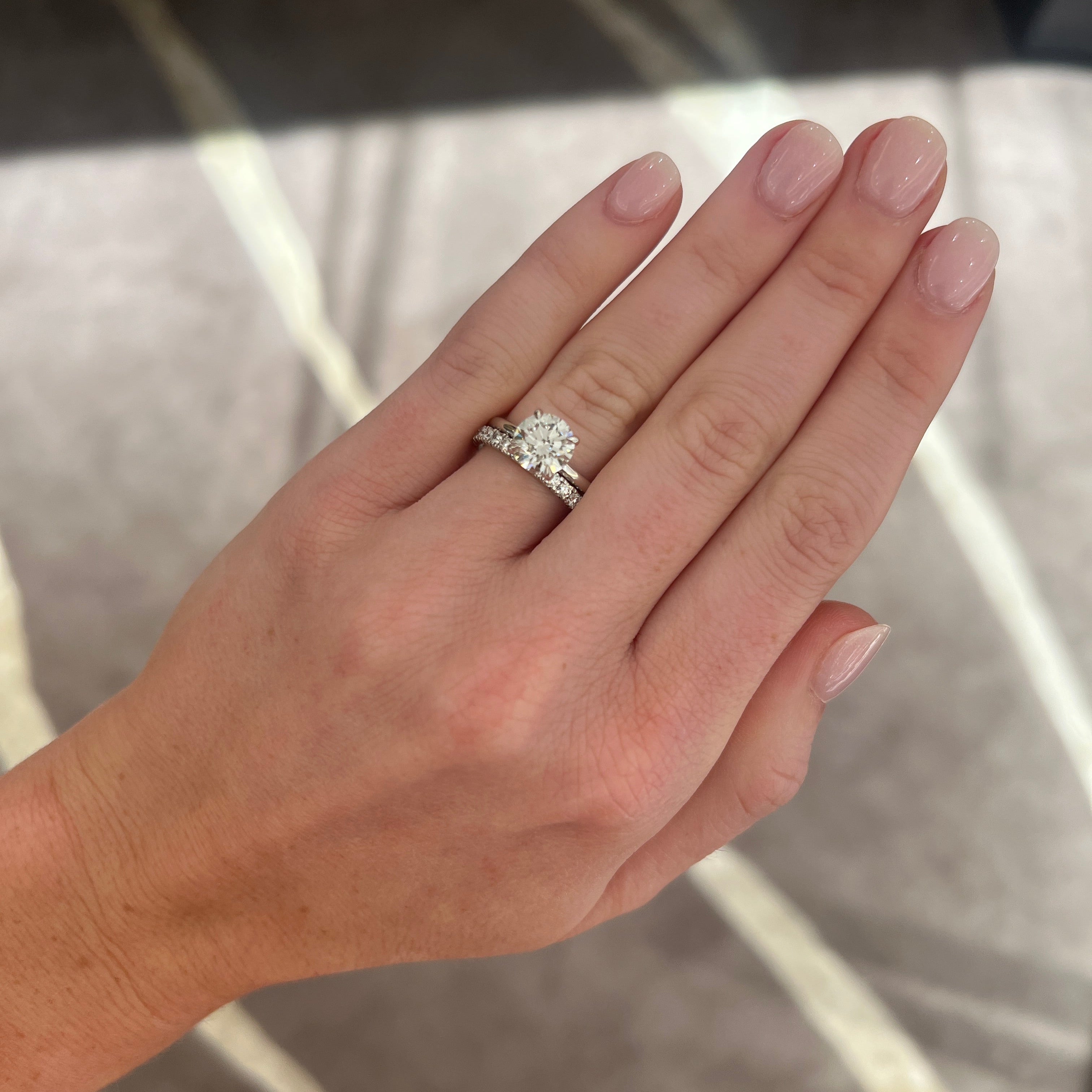 A close-up of a hand wearing a silver-toned, white gold engagement ring with a round-cut diamond, and a round diamond band in white gold, set against a blurred indoor background.