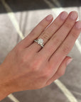 A close-up of a hand wearing a silver-toned, white gold engagement ring with a round-cut diamond, and a round diamond band in white gold, set against a blurred indoor background.