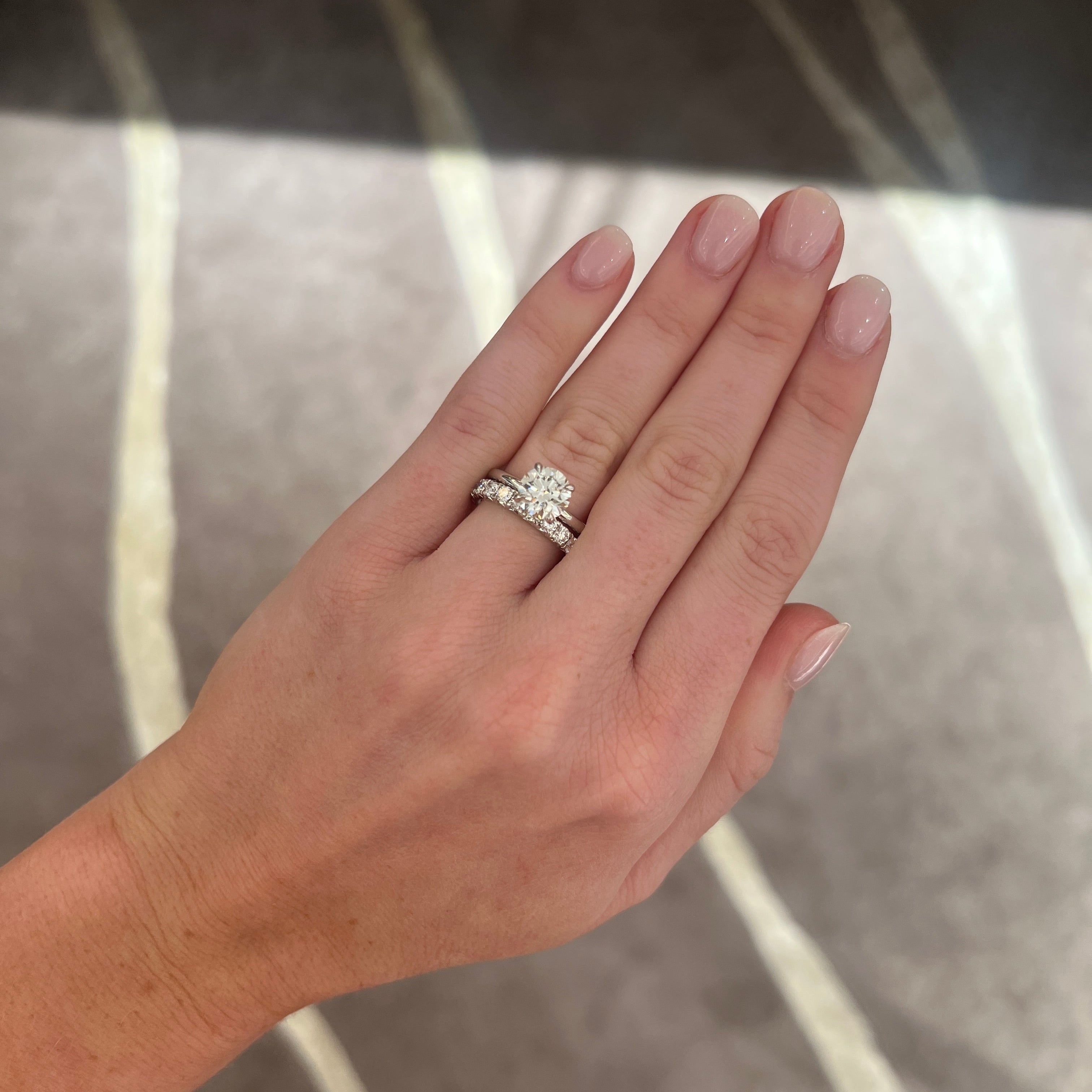 A close-up of a hand wearing a a round diamond solitaire ring, paired with a medium sized platinum band with round diamonds, set against a gray patterned carpet.