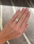A close-up of a hand wearing a a round diamond solitaire ring, paired with a medium sized platinum band with round diamonds, set against a gray patterned carpet.