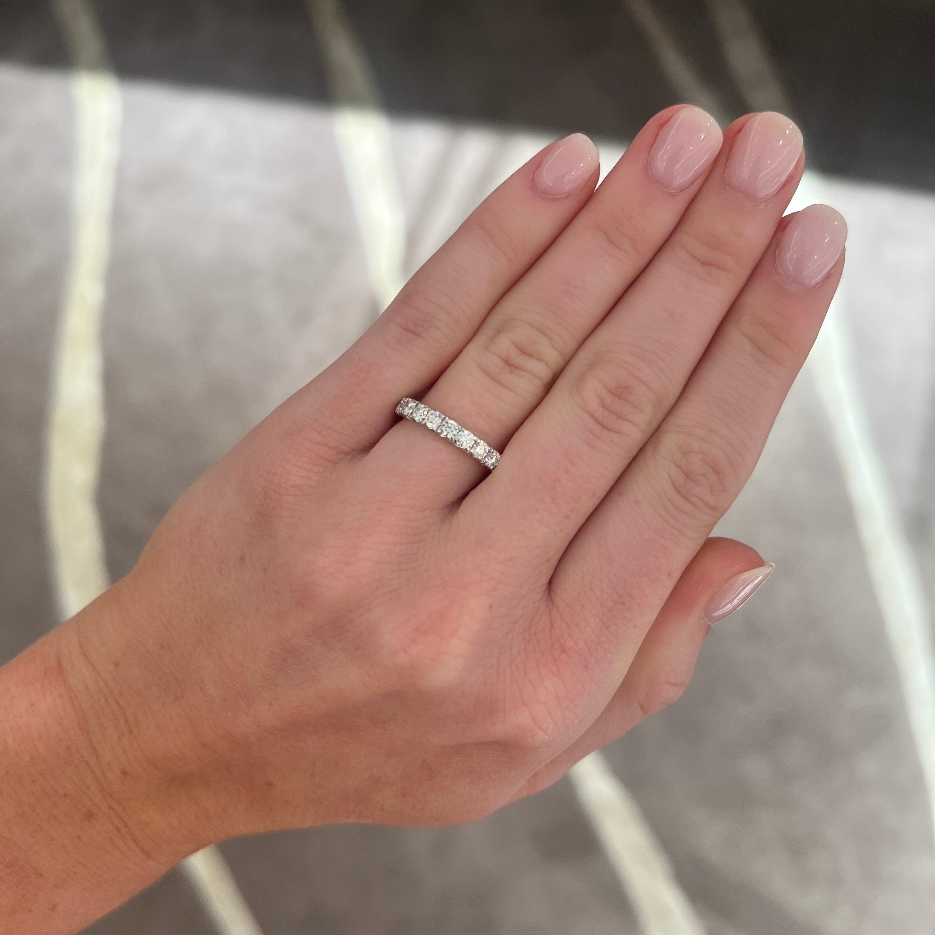A close-up of a hand wearing a medium sized platinum band with round diamonds, set against a gray patterned carpet.