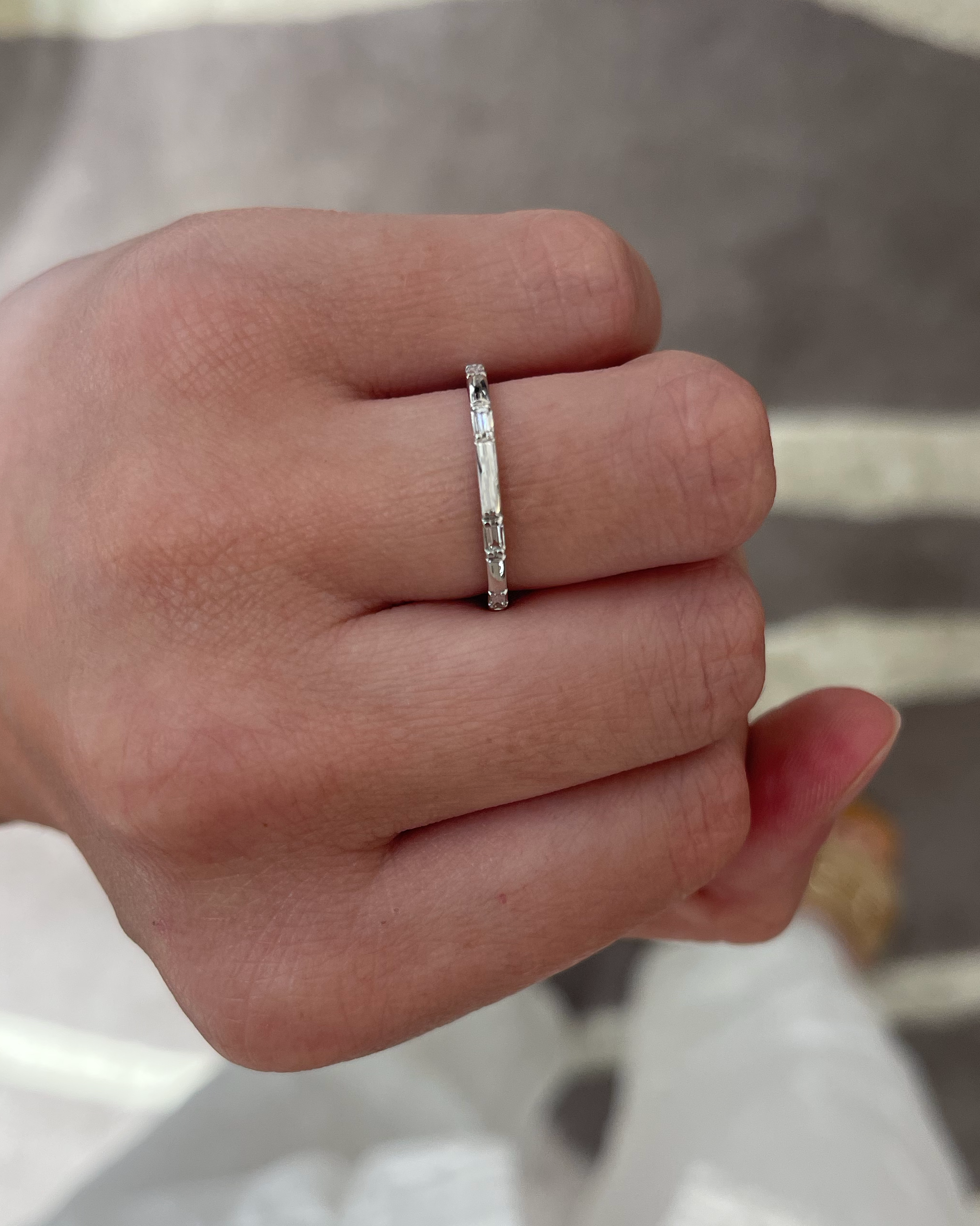 A close-up of a hand wearing a thin silver band ring with small clear stones set along the band, photographed against a blurred gray and white striped background.