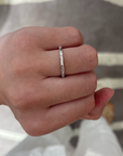A close-up of a hand wearing a thin silver band ring with small clear stones set along the band, photographed against a blurred gray and white striped background.