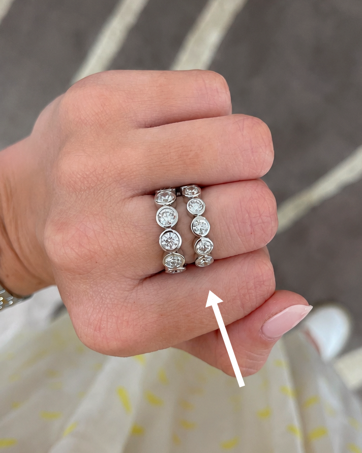 A close-up of a hand wearing two silver rings set with clear round gemstones, with a white arrow pointing to the rings. The background shows a patterned gray rug and a light-colored dress with yellow dots.