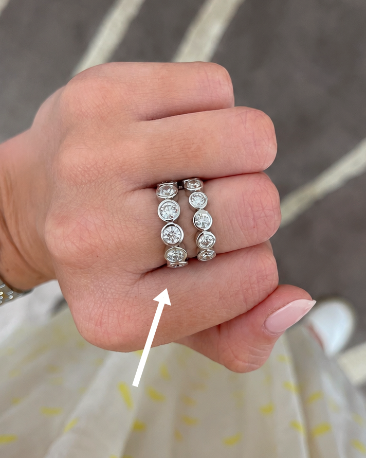 A close-up of a hand wearing two silver rings set with round clear gemstones, with a white arrow pointing to the rings. The background shows a patterned gray rug and a light-colored dress with yellow spots.