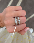 A close-up of a hand wearing two silver rings set with round clear gemstones, with a white arrow pointing to the rings. The background shows a patterned gray rug and a light-colored dress with yellow spots.