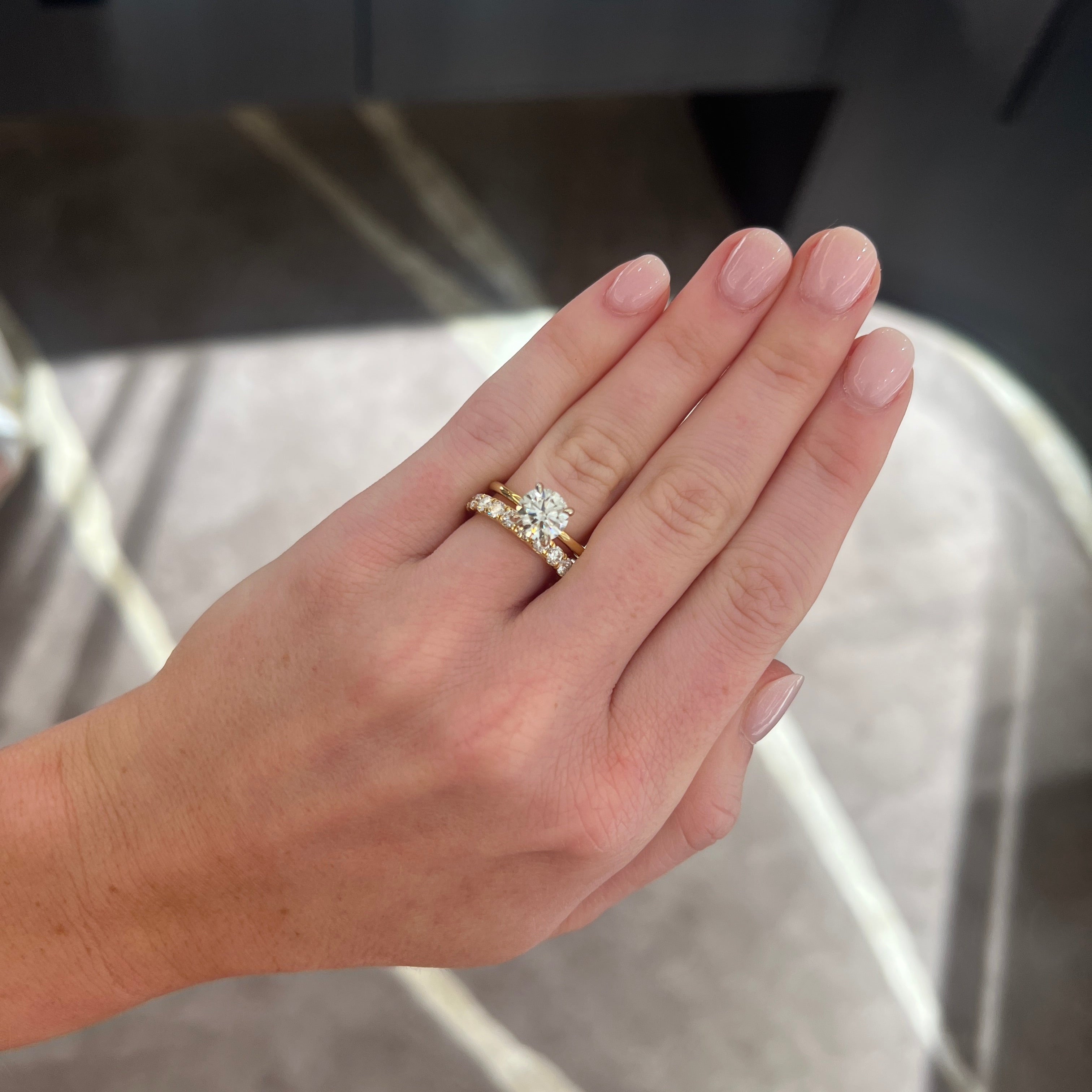 A close-up of a hand wearing a round diamond solitaire engagement ring paired with a medium sized 1.02 carat yellow gold wedding band ring with round diamonds, set against a blurred gray and white marble-patterned background.