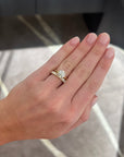 A close-up of a hand wearing a round diamond solitaire engagement ring paired with a medium sized 1.02 carat yellow gold wedding band ring with round diamonds, set against a blurred gray and white marble-patterned background.