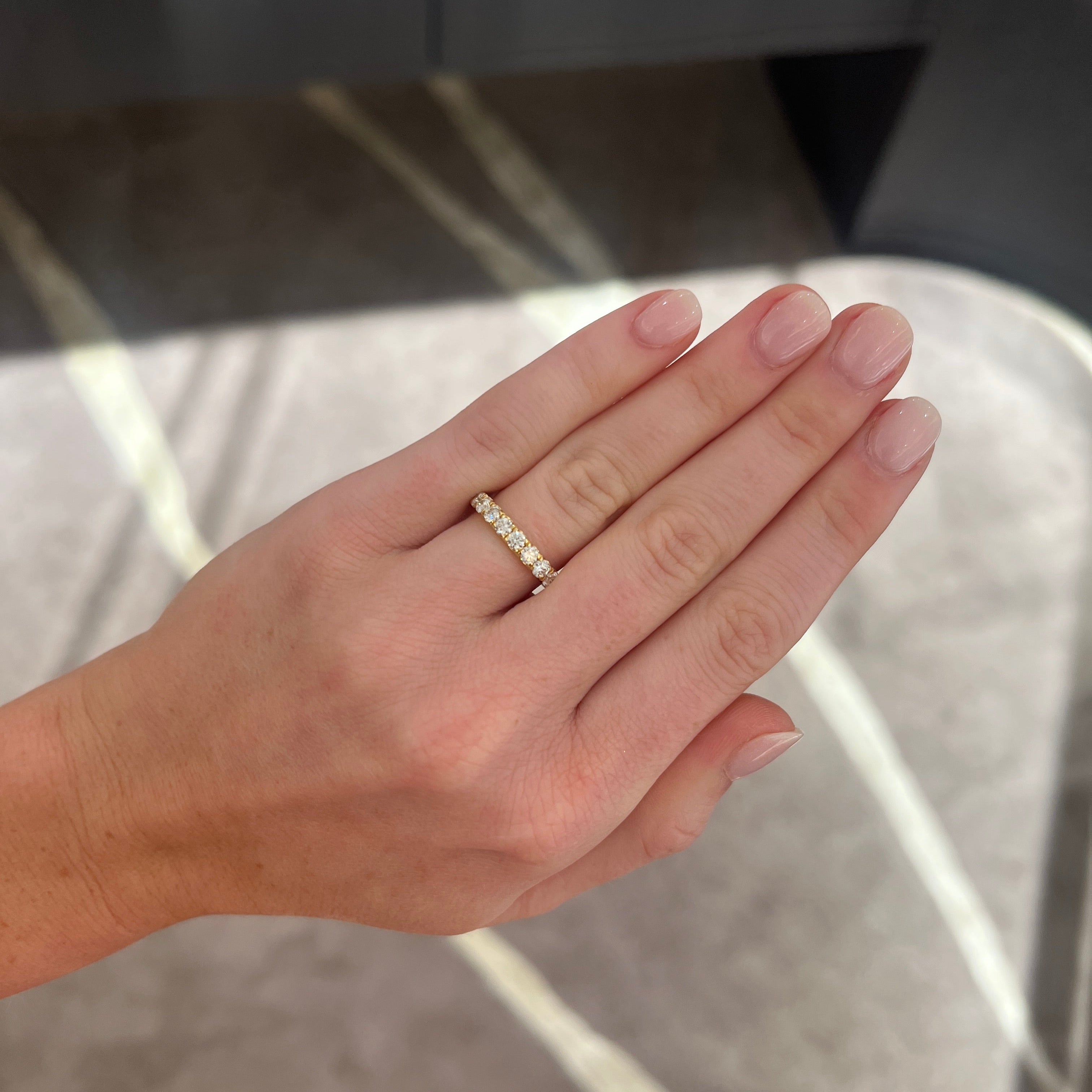 A close-up of a hand wearing a medium sized 1.02 carat yellow gold wedding band ring with round diamonds, set against a blurred gray and white marble-patterned background.