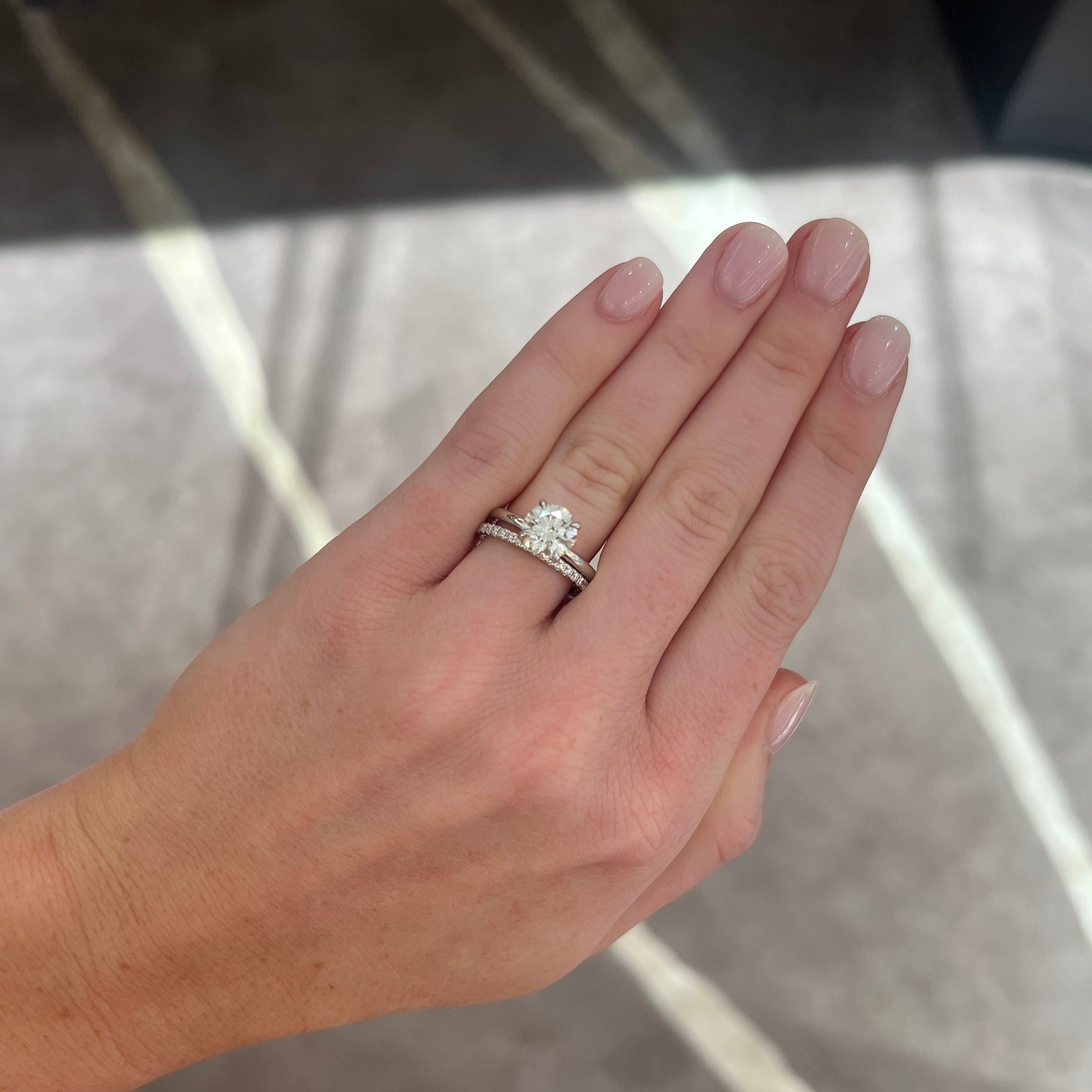 A close-up of a hand wearing a white gold engagement ring with a round-cut diamond and a matching band set with small diamonds, shown against a gray patterned floor background.