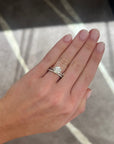 A close-up of a hand wearing a white gold engagement ring with a round-cut diamond and a matching band set with small diamonds, shown against a gray patterned floor background.
