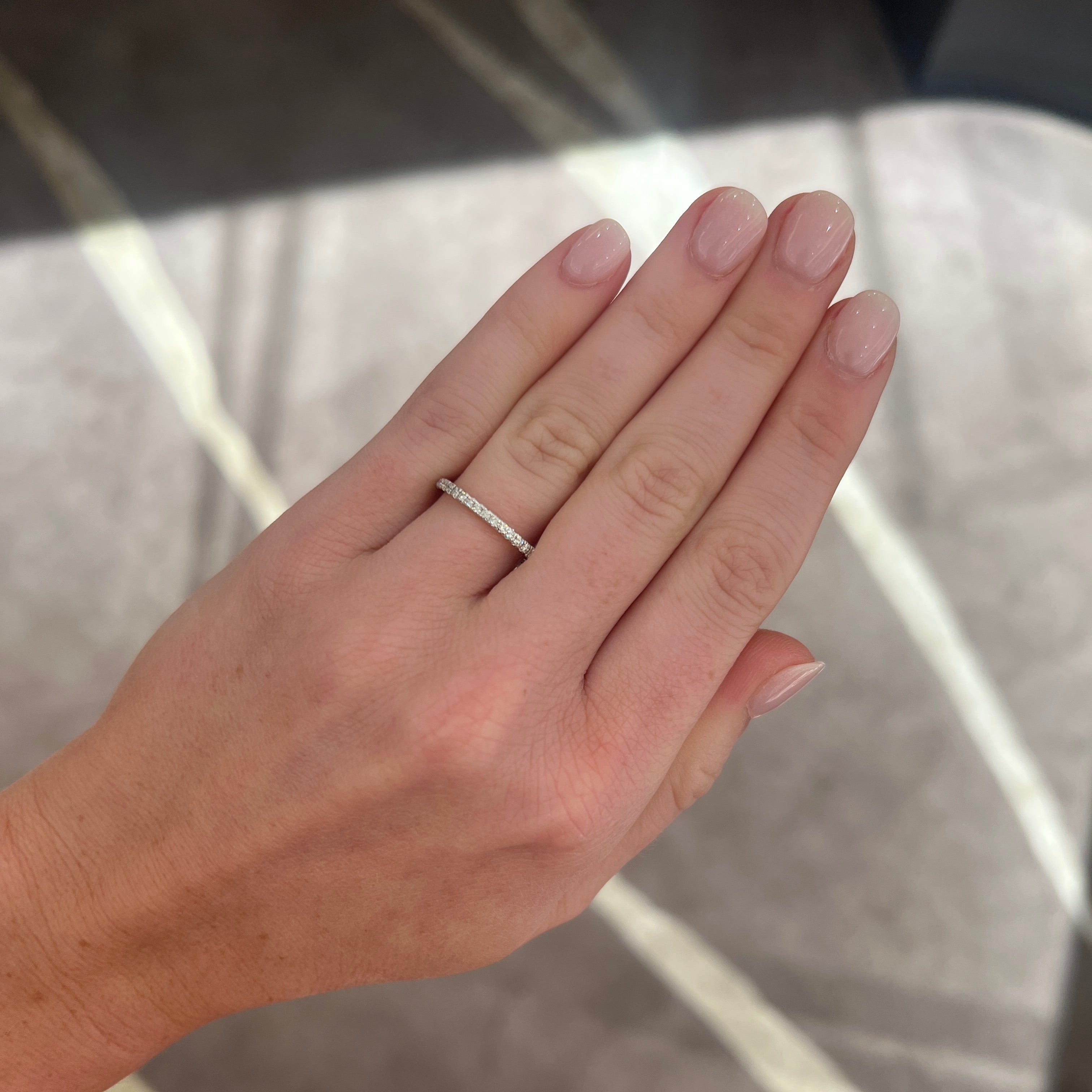 A close-up of a person's hand wearing a slim white gold diamond band ring set against a gray marble-patterned background.