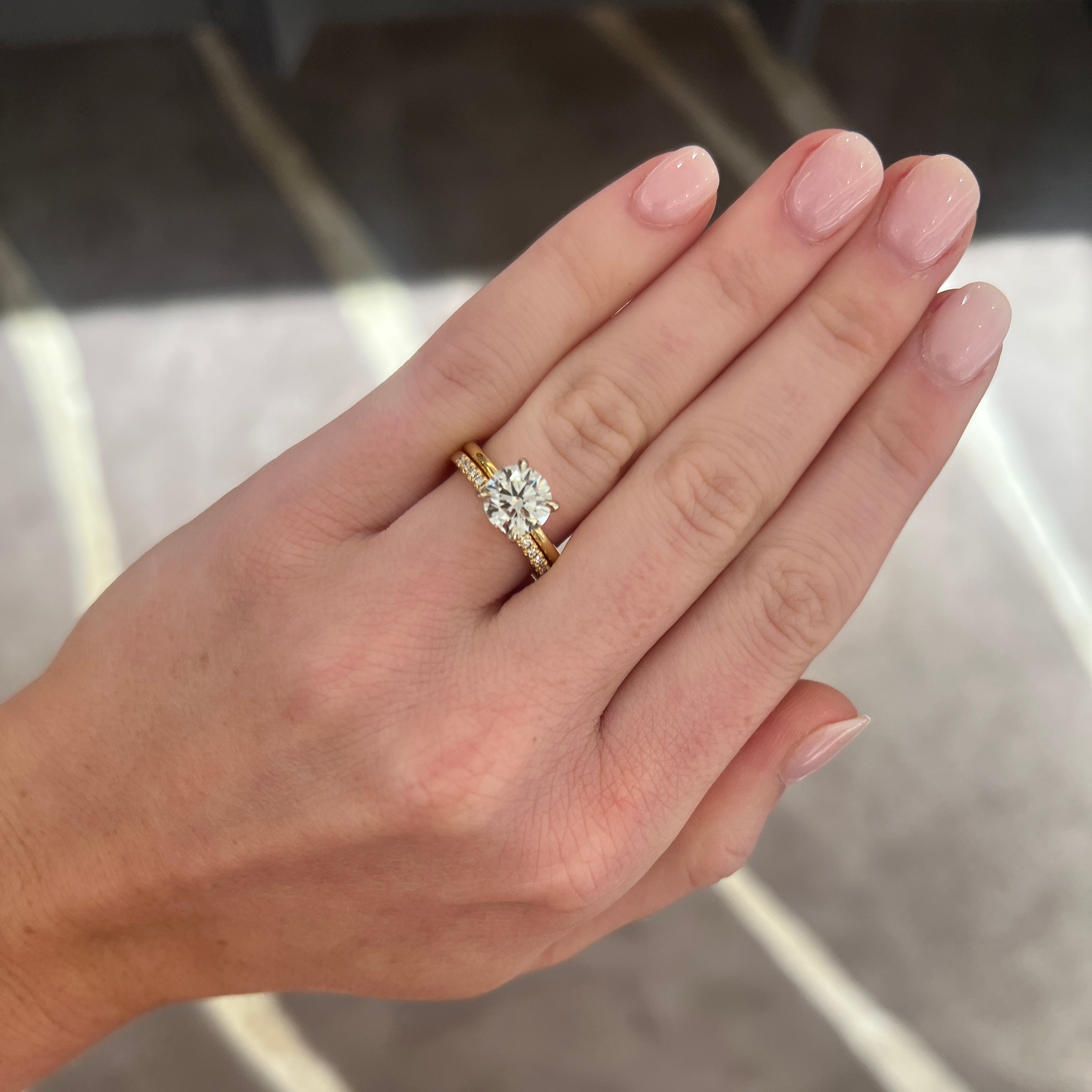 A close-up of a hand wearing a yellow gold ring with a large round-cut diamond cluster, set against a blurred gray patterned background.