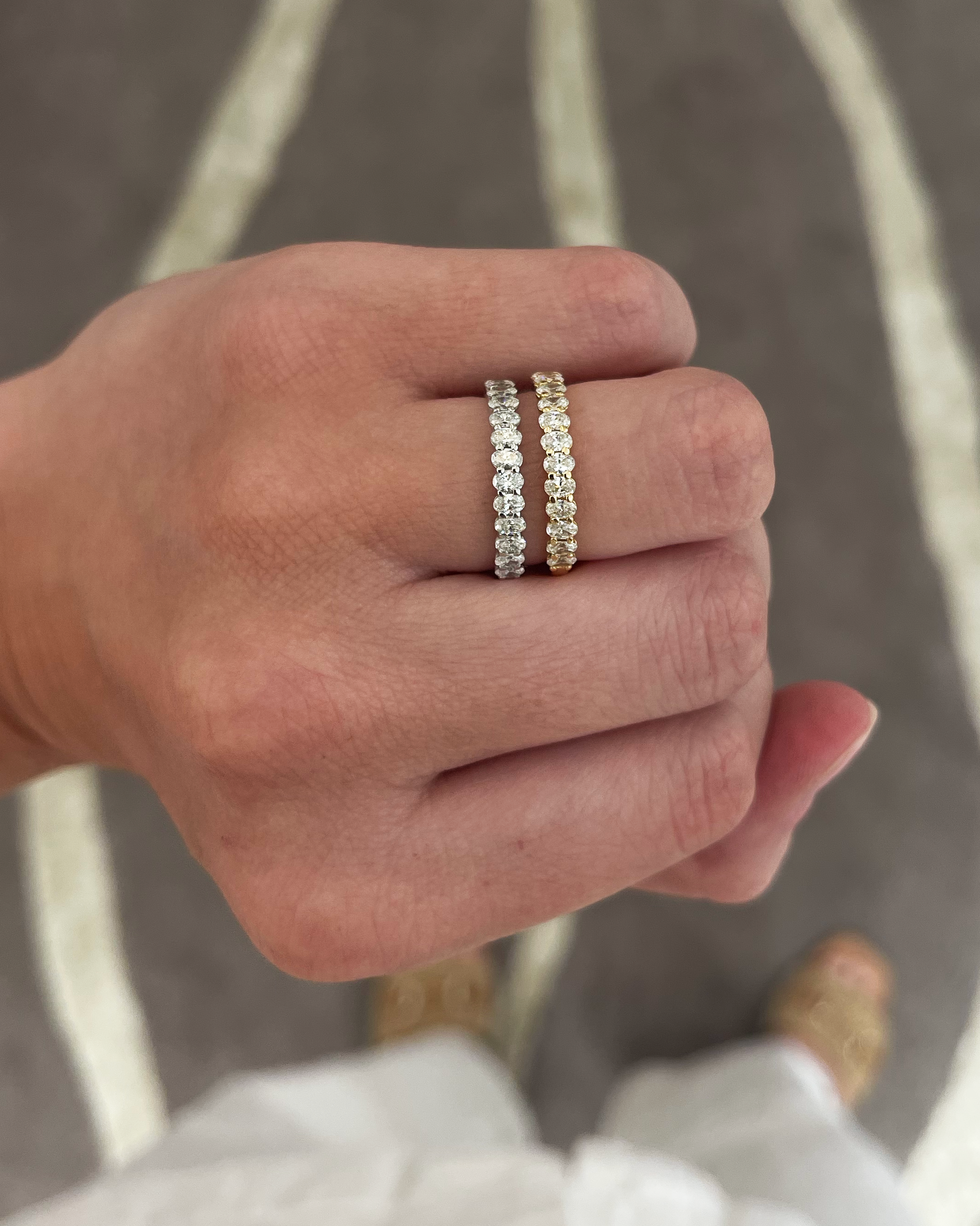 A close-up of a person's hand wearing two slim eternity rings. One ring is silver with clear stones, and the other is yellow gold with clear stones, set against a gray and white patterned rug.