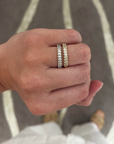 A close-up of a person's hand wearing two slim eternity rings. One ring is silver with clear stones, and the other is yellow gold with clear stones, set against a gray and white patterned rug.