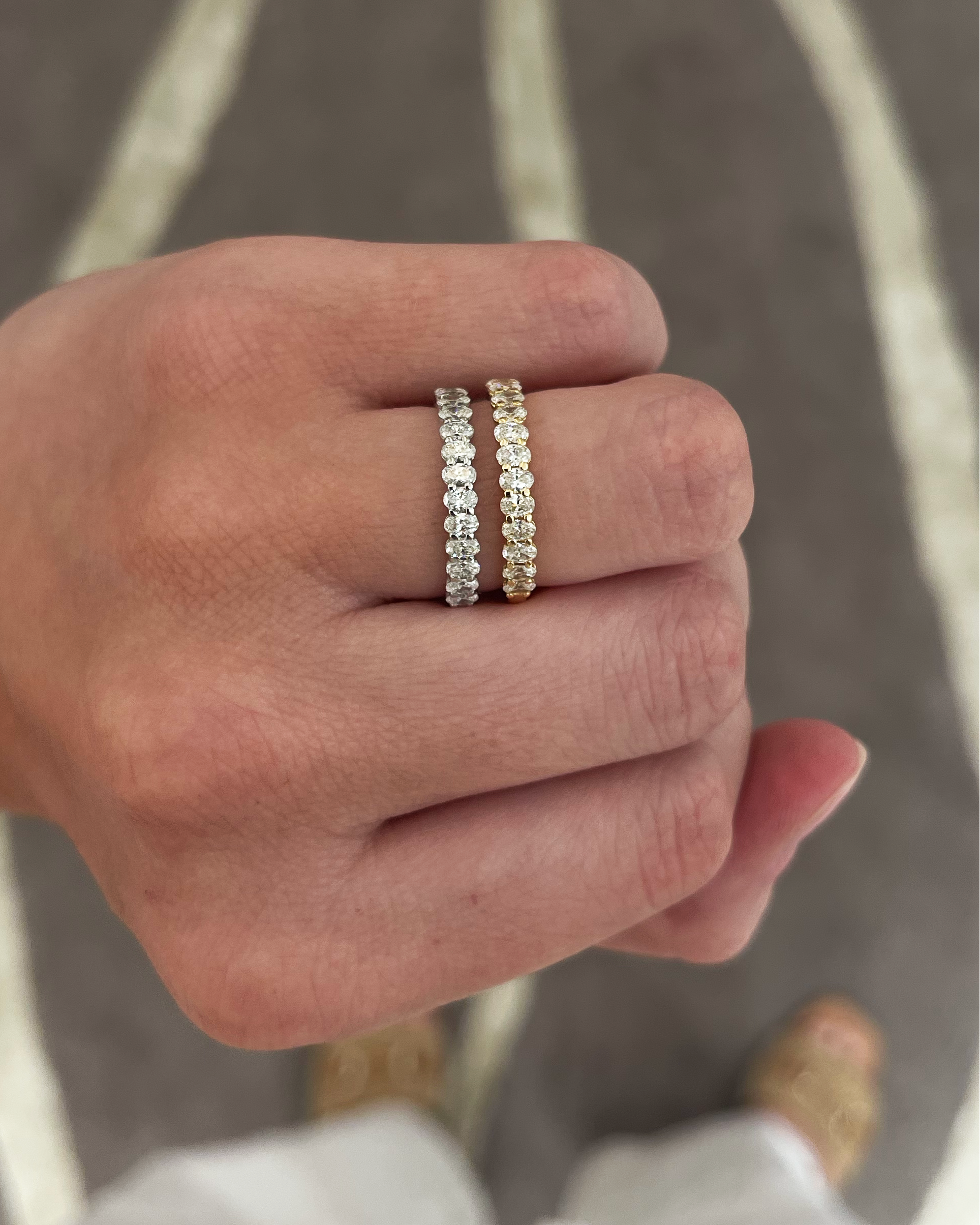 A close-up of a person's hand wearing two slim eternity rings. One ring is silver with clear stones, and the other is yellow gold with clear stones, set against a gray and white patterned rug.
