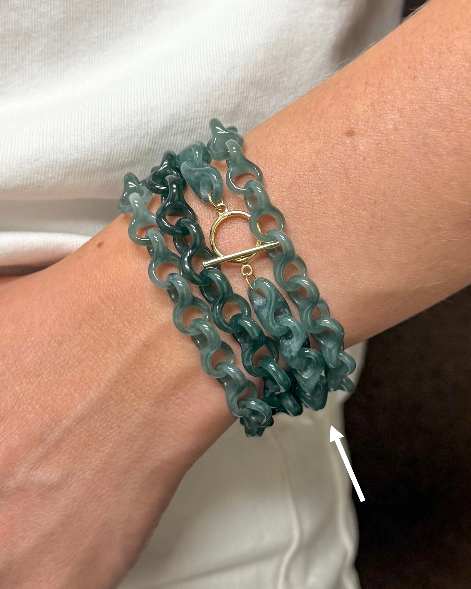 A close-up of a person's wrist wearing a multi-strand teal chain bracelet with a gold toggle clasp, against a white shirt background.