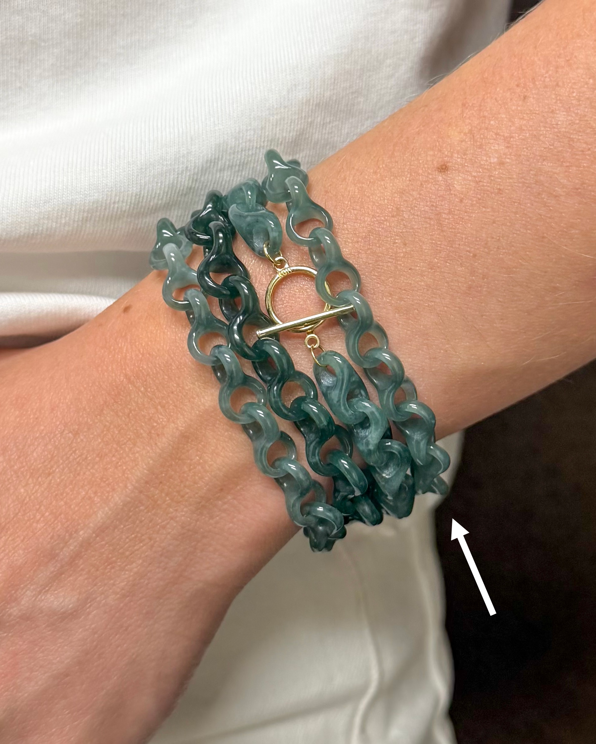 A close-up of a person's wrist wearing a multi-strand teal-green chain bracelet with a gold toggle clasp, against a white shirt background.