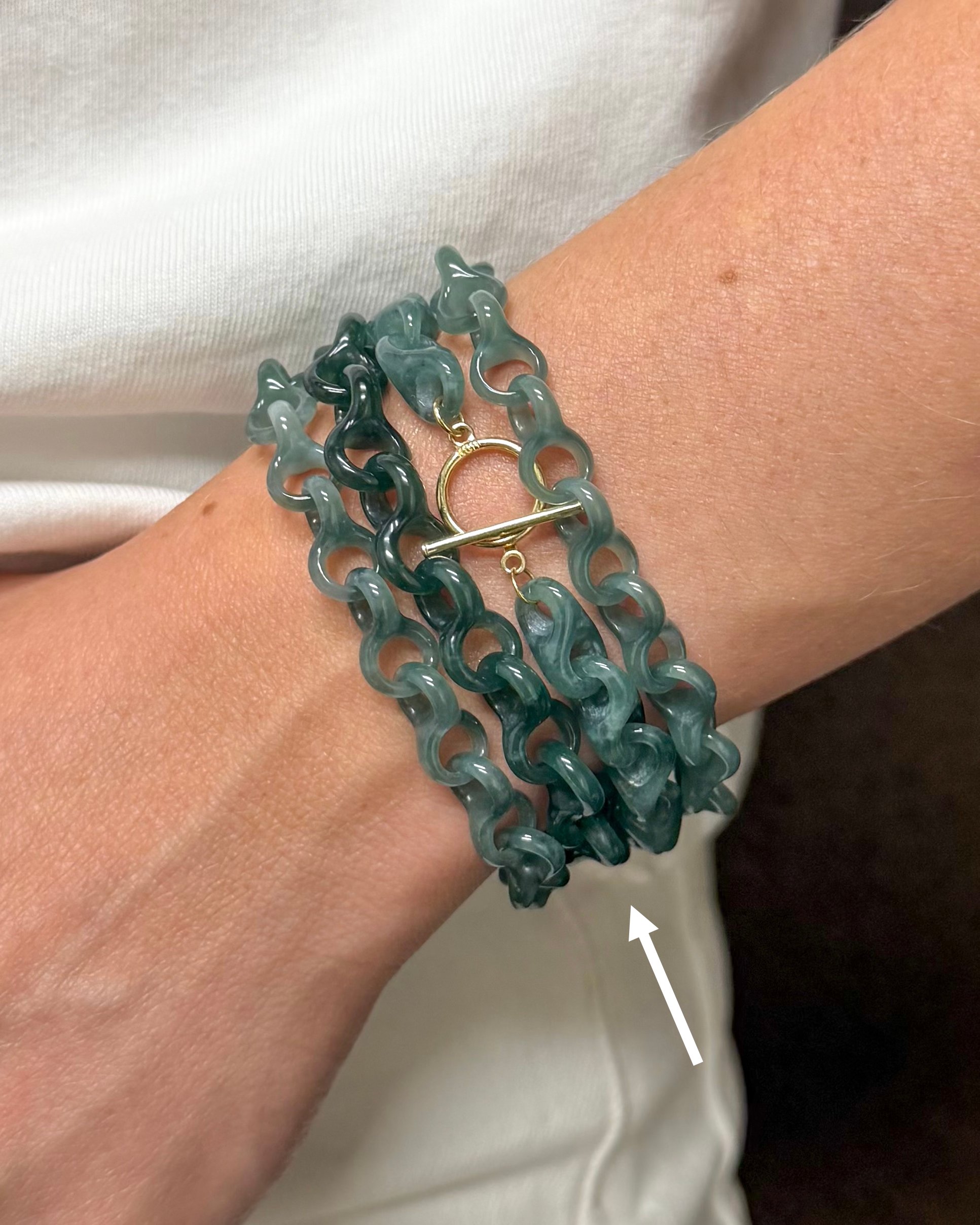 A close-up of a person's wrist wearing a multi-strand teal chain bracelet with a gold toggle clasp, with a white arrow pointing to the bracelet.