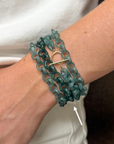 A close-up of a person's wrist wearing a multi-strand teal chain bracelet with a gold toggle clasp, with a white arrow pointing to the bracelet.
